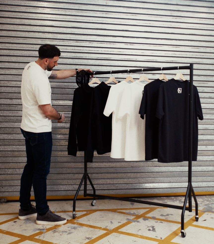 Man hanging a cap on a clothing rail, the clothing rail has a mixture of white and black 'OFFEND' T-shirts and 'OFFEND' Hoodie on. The back ground is a shutter