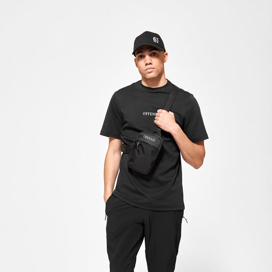Model wearing 'OFFEND' mesh trucker cap, 'OFFEND ' black T-Shirt with 'OFFEND' text across centre of chest, wearing classic black 'OFFEND' crossbody bag with 'OFFEND' text across top of bag