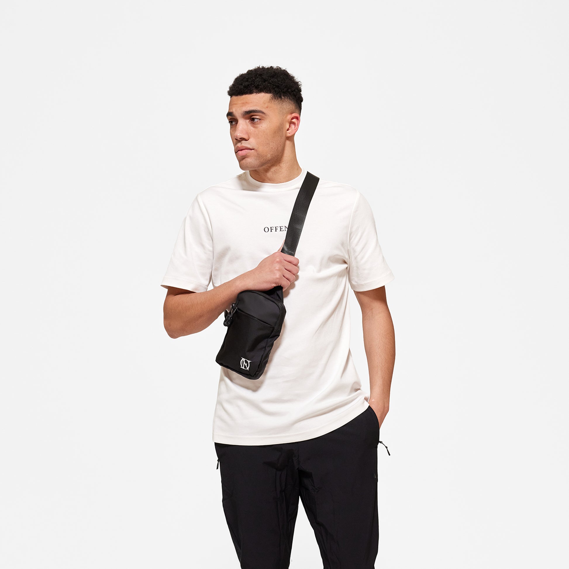 Model wearing white 'OFFEND' T-shirt with black 'OFFEND' text across centre of chest, wearing black crossbody bag with white 'OFFEND' logo in the bottom right hand corner of bag