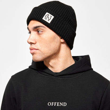 Close up shot of the model wearing black 'OFFEND' beanie with a black 'OFFEND' logo on a white background, and black 'OFFEND' hoodie with 'OFFEND' text across centre of chest 