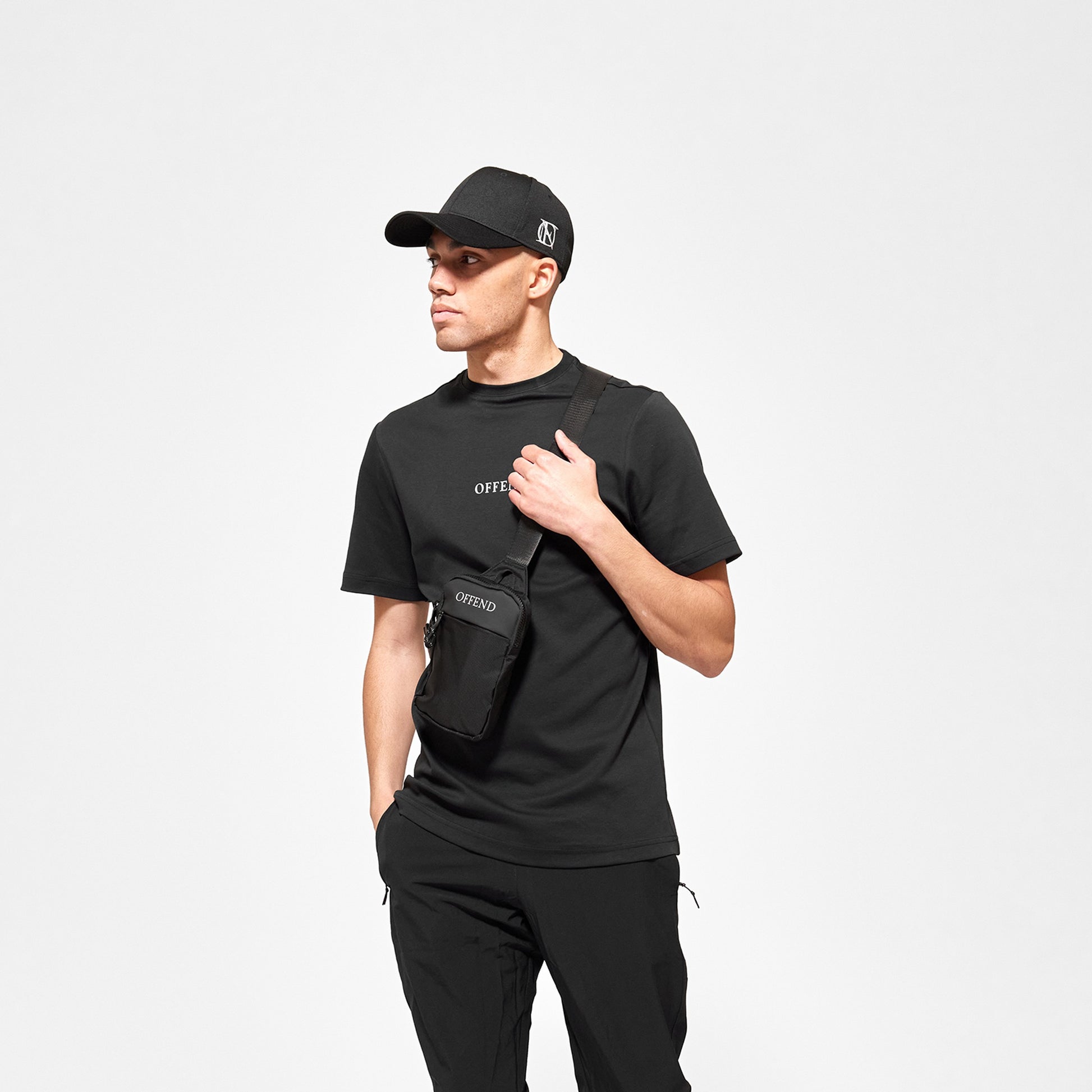 Model wearing 'OFFEND' black T shot from the front with 'OFFEND' text on chest wearing the classic 'OFFEND' cross body bag, wearing 'OFFEND' baseball cap
